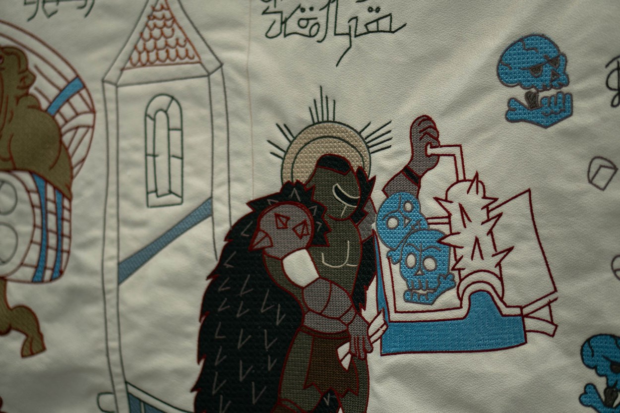 Close-up of a section of the Slayeux Tapestry showing the DOOM Slayer in medieval-style embroidery, holding a shield with demonic skulls and surrounded by stylized symbols and gothic architecture.