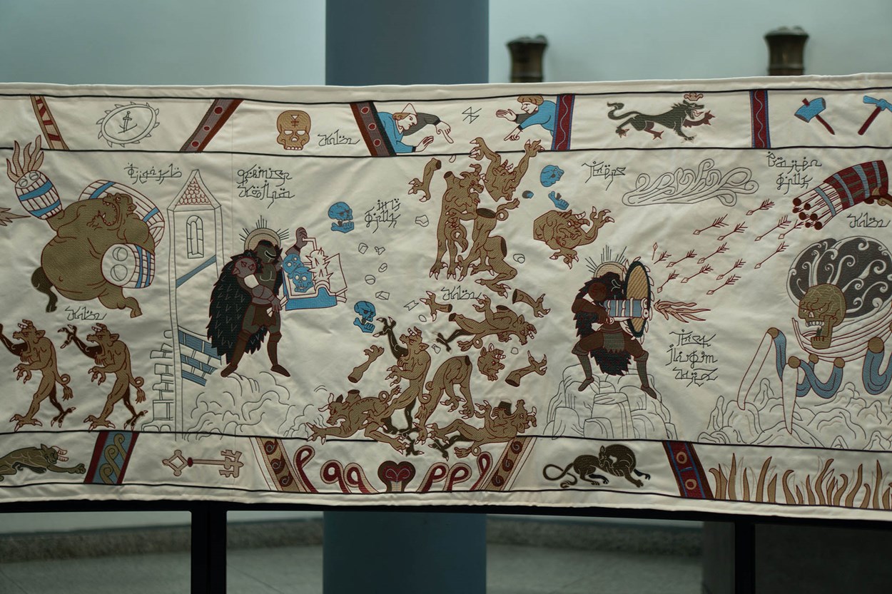 A section of the Slayeux Tapestry, styled after the Bayeux Tapestry, depicting the DOOM Slayer battling demons with medieval-inspired artwork. The embroidery features monsters, weaponry, and symbolic imagery in rich brown, red, and blue tones on a cream background.