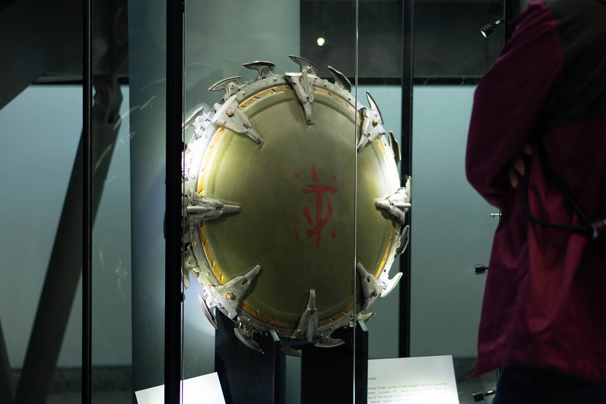 The full-scale, functional Shield Saw prop from DOOM: The Dark Ages on display in a glass case at the Royal Armouries Museum, with a visitor observing it up close.