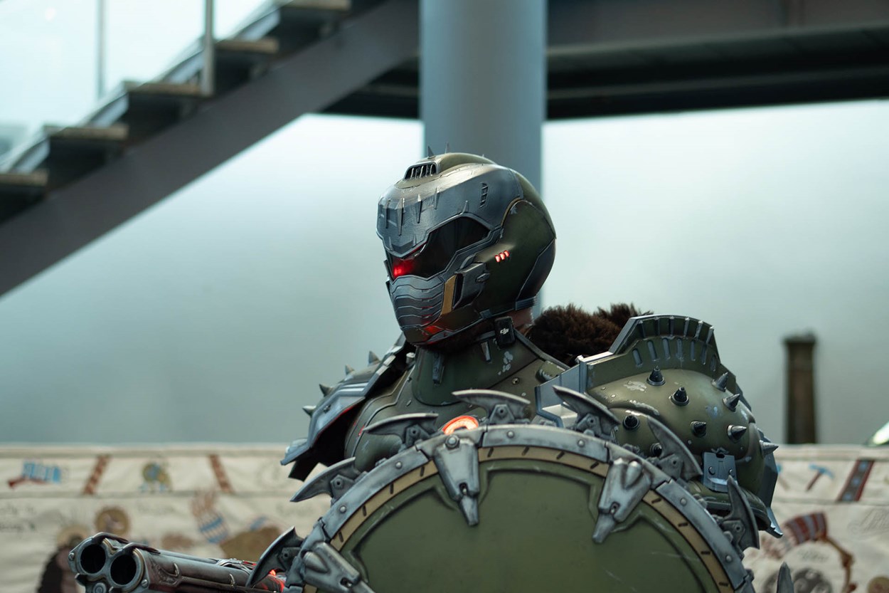 Close-up of Olivier Richters in full DOOM Slayer armor, holding the Shield Saw and a double-barrel shotgun prop, with detailed spiked armor and glowing red helmet visor at the Royal Armouries event.