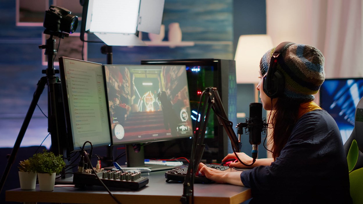 A female gamer wearing a beanie and headset is live streaming from her gaming setup. She is playing a game on a dual-monitor setup, with one screen displaying gameplay and the other showing a chat window. A professional microphone, audio mixer, and camera setup are visible, along with soft studio lighting, creating an immersive streaming environment.