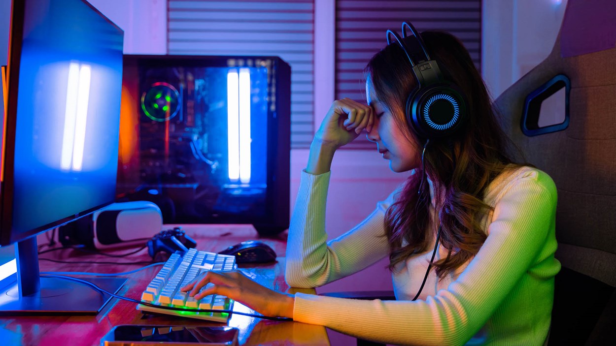 A young female gamer wearing a headset sits at a gaming desk with RGB lighting, resting her head on her hand in frustration. A dual-monitor setup and gaming PC with LED lights are visible in the background.