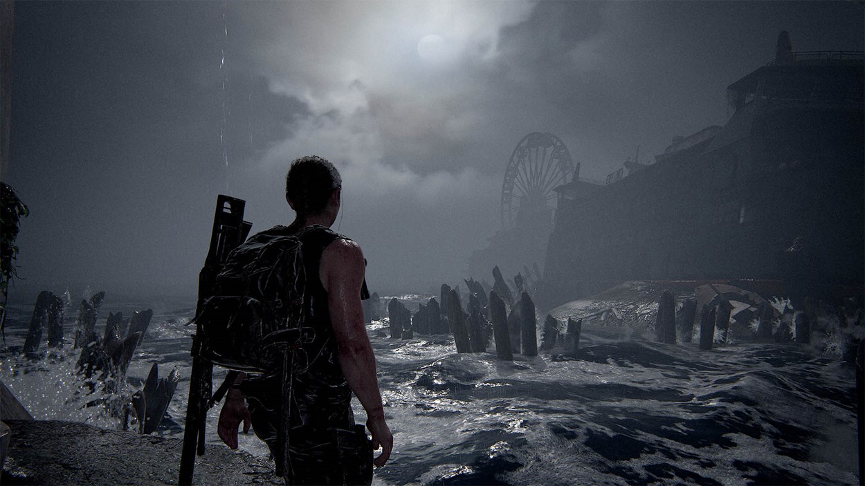 A lone character stands at the edge of a stormy shoreline in The Last of Us Part II Remastered, staring toward a ruined pier and a distant ferris wheel under a cloudy, moonlit sky.