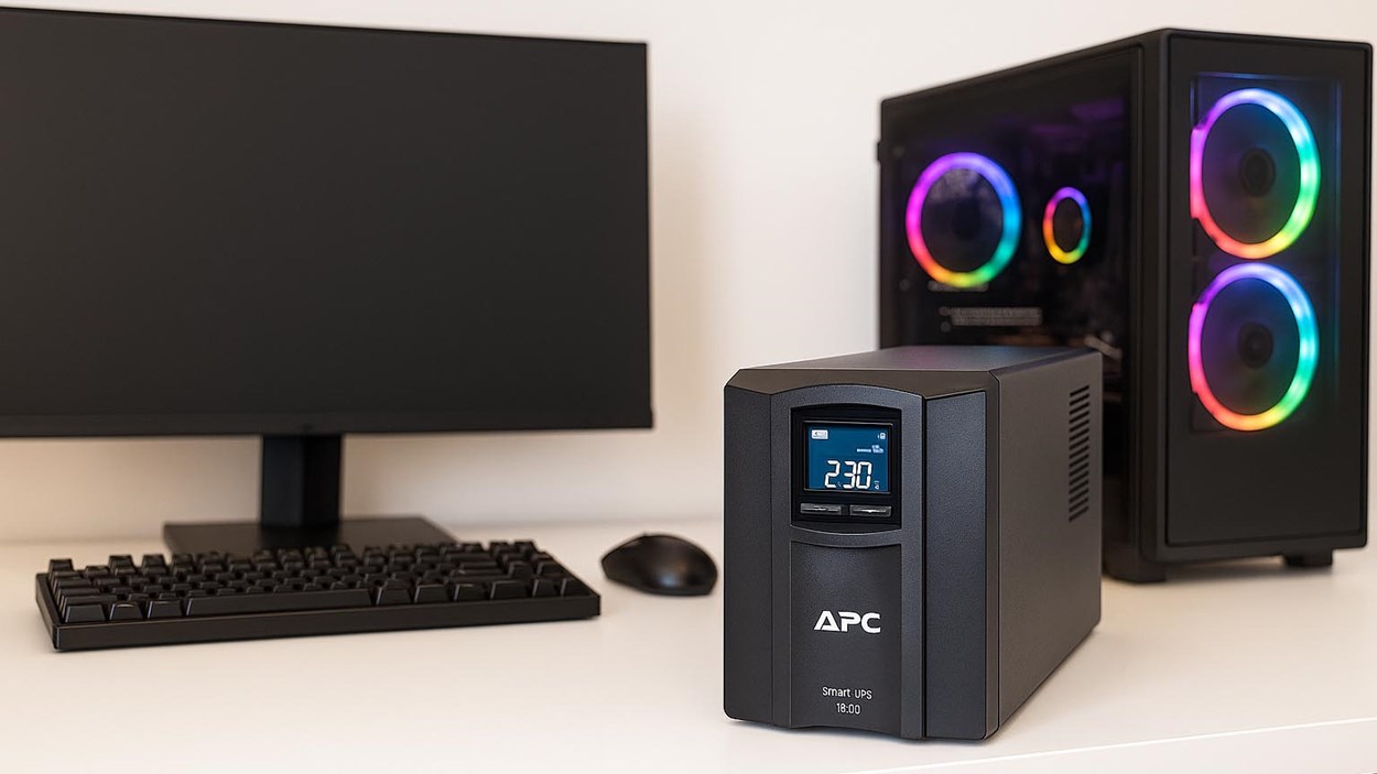 A sleek desktop gaming setup featuring an APC Smart-UPS 1000 with a digital display, placed next to a modern RGB-lit gaming PC, widescreen monitor, mechanical keyboard, and mouse on a white desk