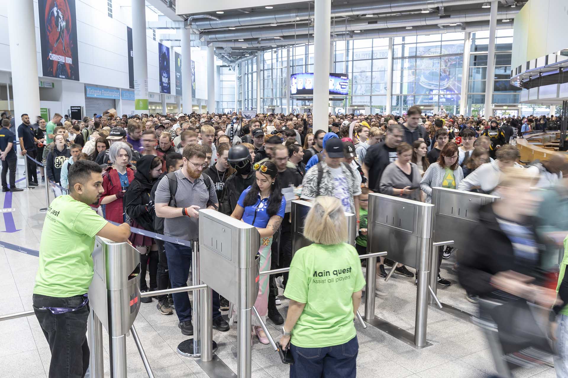 Large crowd of gamers entering Gamescom 2025 through the main gates, guided by staff in green shirts marked 'Main quest: Assist all players'.