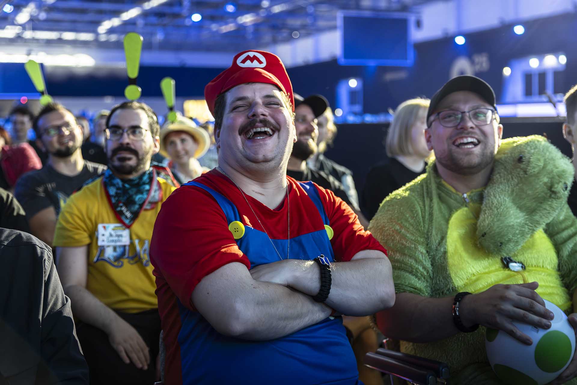 Fans in cosplay at Gamescom 2025, including someone dressed as Mario and another in a green dinosaur outfit holding a Yoshi egg, enjoying the show.