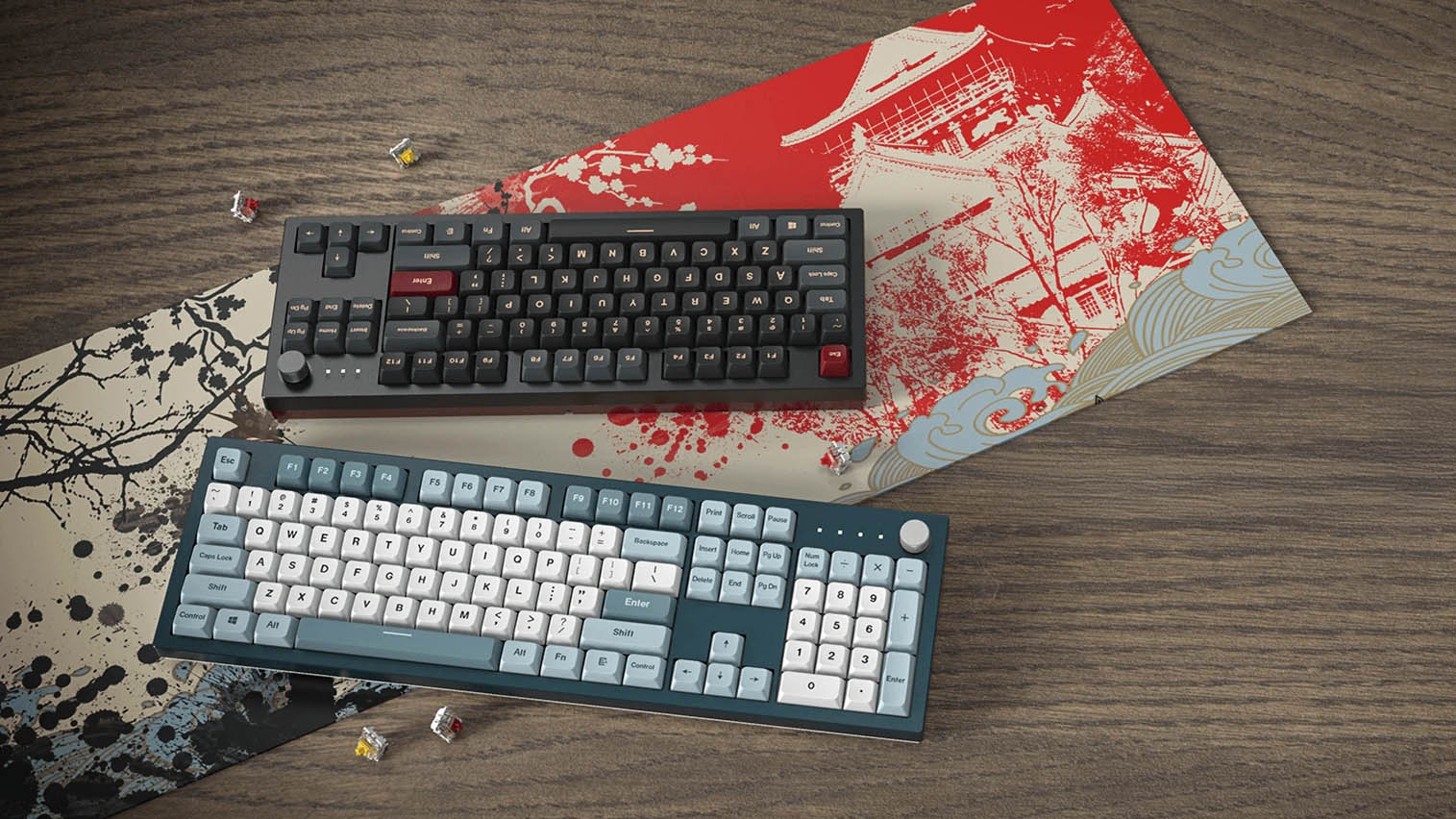 Two Montech MKey mechanical keyboards placed on Japanese art-inspired gaming mouse pads. One keyboard has a dark-themed design with red and black keys, while the other features a light blue and white color scheme. Loose mechanical switches are scattered around on a wooden surface.