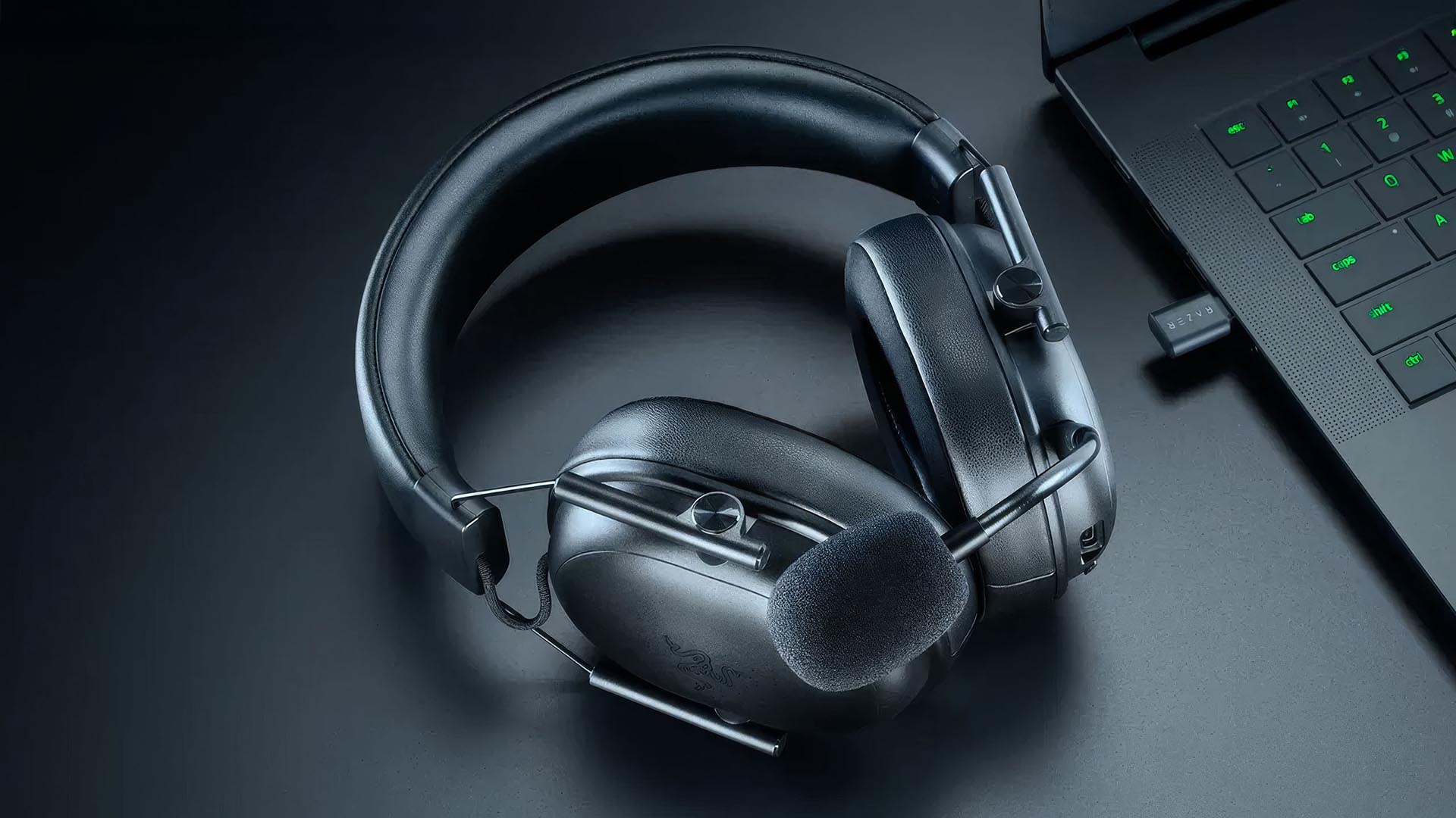 A Razer BlackShark V3 X HyperSpeed wireless gaming headset with boom microphone resting beside a Razer laptop on a dark desk.