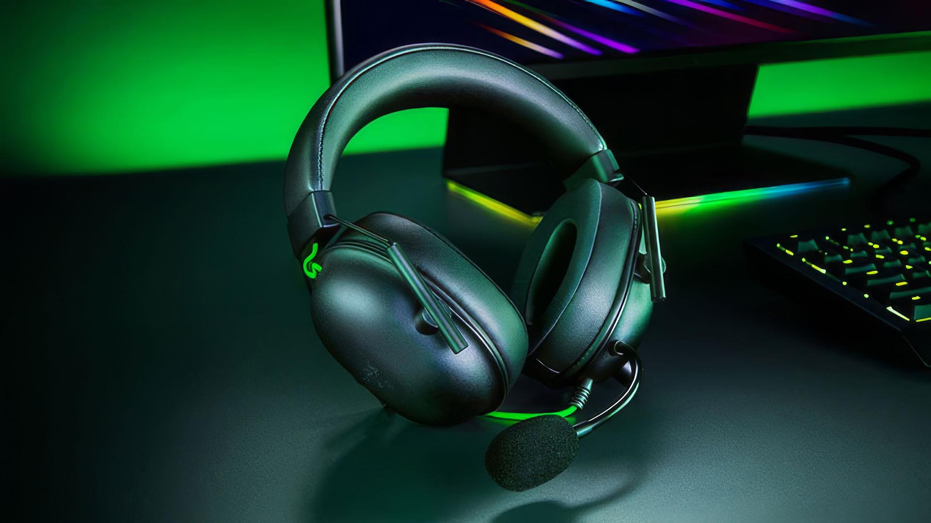 A Razer BlackShark V2 X gaming headset resting on a desk with green lighting and a RGB keyboard in the background.