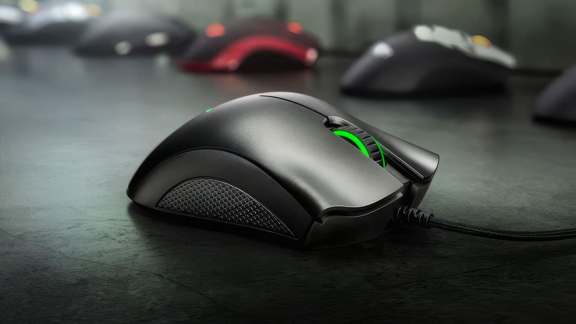 A close up of a Razer DeathAdder gaming mouse on a desk surface with its green scroll wheel illuminated and other mice blurred in the background.