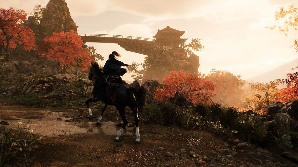 A samurai rides a horse through a picturesque autumn landscape, surrounded by vibrant red and orange foliage. In the distance, a traditional Japanese pagoda-style building sits atop a rocky cliff, connected by an elegant wooden bridge. The golden light of the setting sun bathes the scene, enhancing the tranquil yet adventurous atmosphere of Rise of the Ronin.