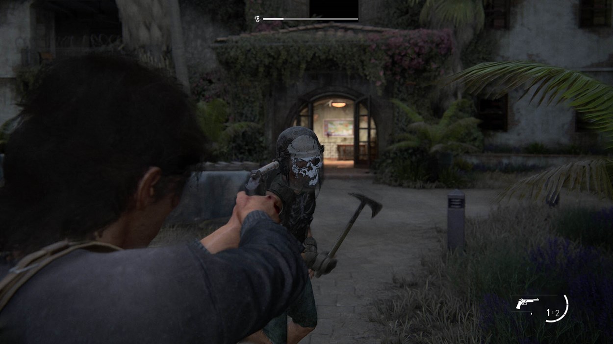 Ellie aims a pistol at a hostile enemy wearing a skull-painted helmet and wielding a pickaxe in The Last of Us Part II Remastered, set outside a lush, overgrown compound.